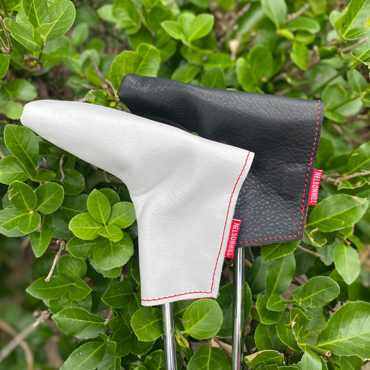 Two golf putter headcovers, one white and one black textured leather, both with red stitching and red brand tags, shown outdoors against a background of green leaves.