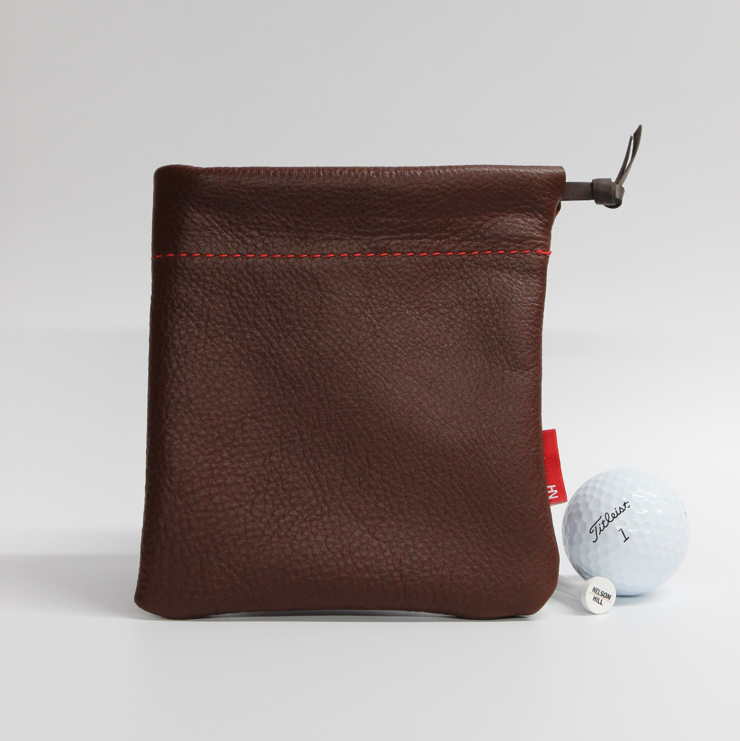 Brown leather pouch with red stitching on a white background, featuring a golf ball and club head cover.