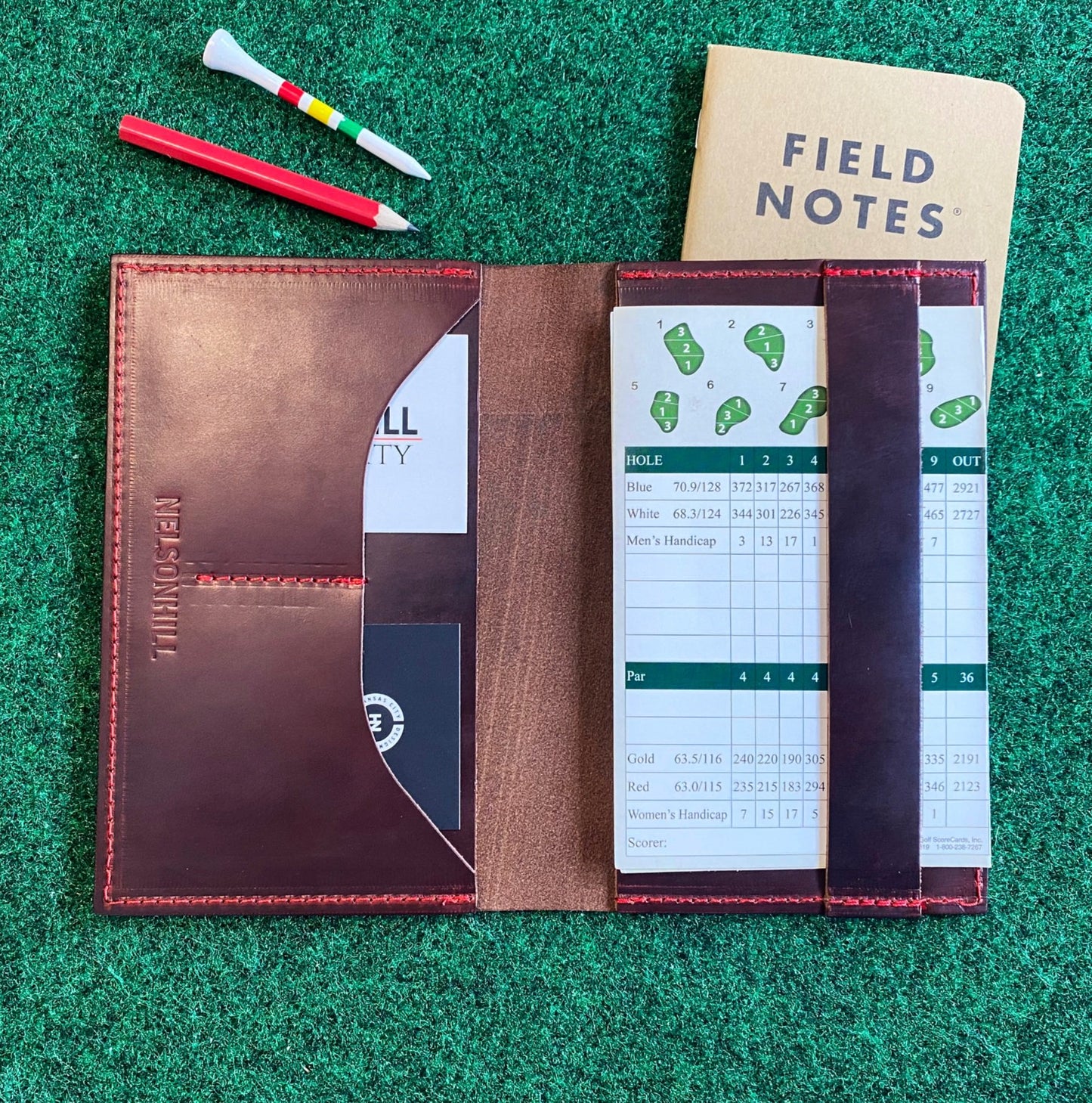 Brown leather notebook with a white scorecard on a green grass-like surface, featuring 'Field Notes' journal.