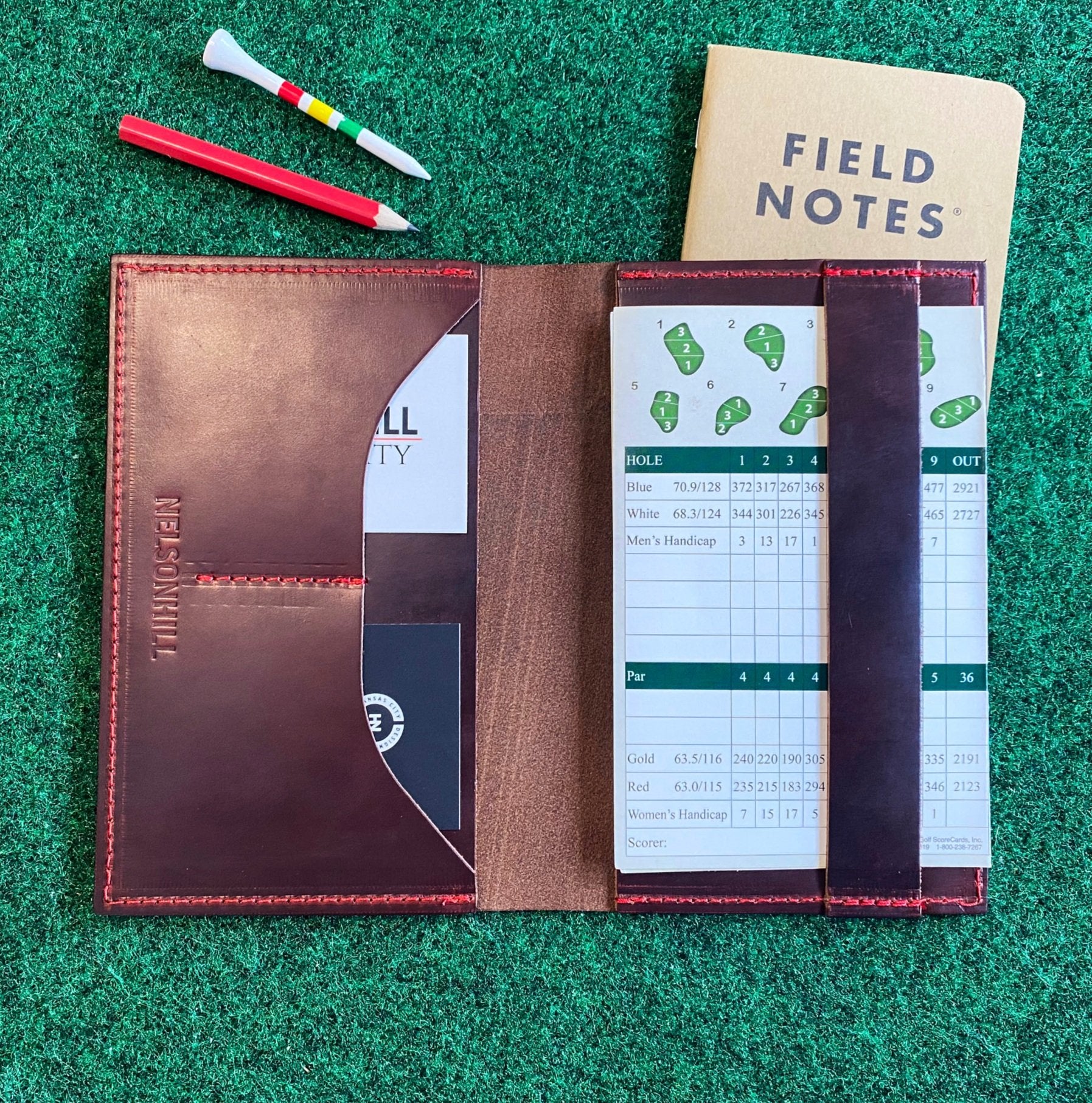 Brown leather notebook with a white scorecard on a green grass-like surface, featuring 'Field Notes' journal.
