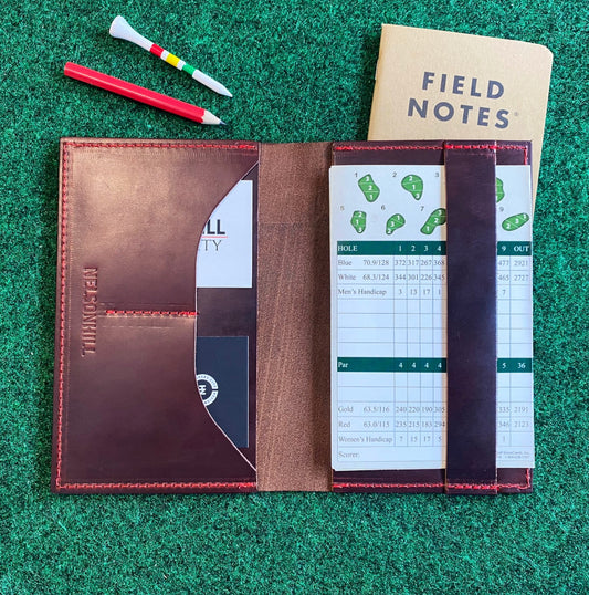 Brown leather notebook with a white scorecard on a green grass-like surface, featuring 'Field Notes' journal.