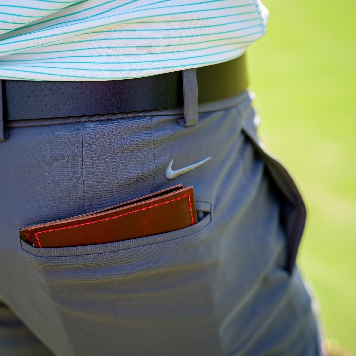 Person wearing a light blue striped shirt and gray pants with a brown golf journal in a pocket with a blurred green background